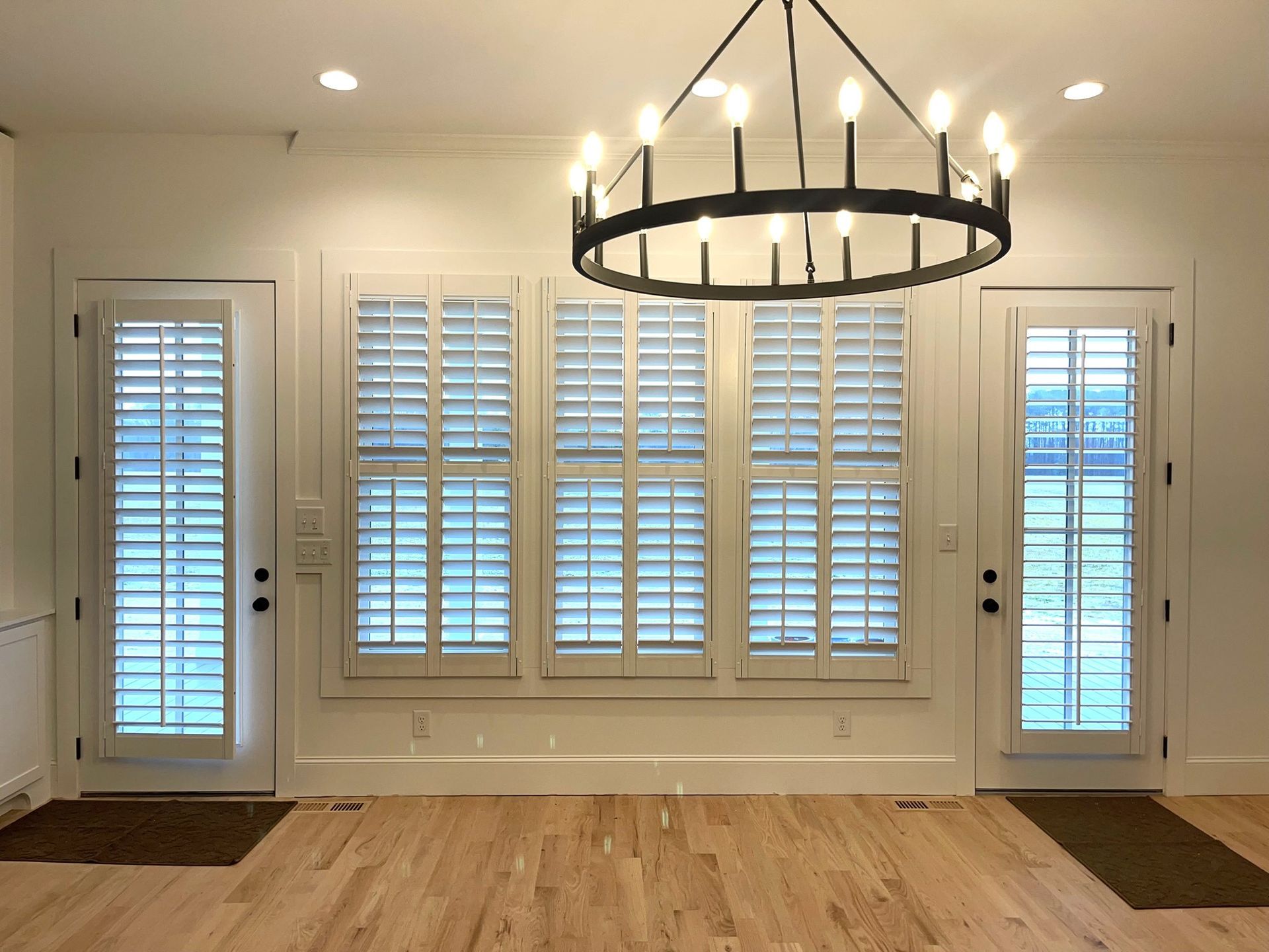 A bright, interior room with white shutters on windows and doors, hardwood floors, and a black chandelier.