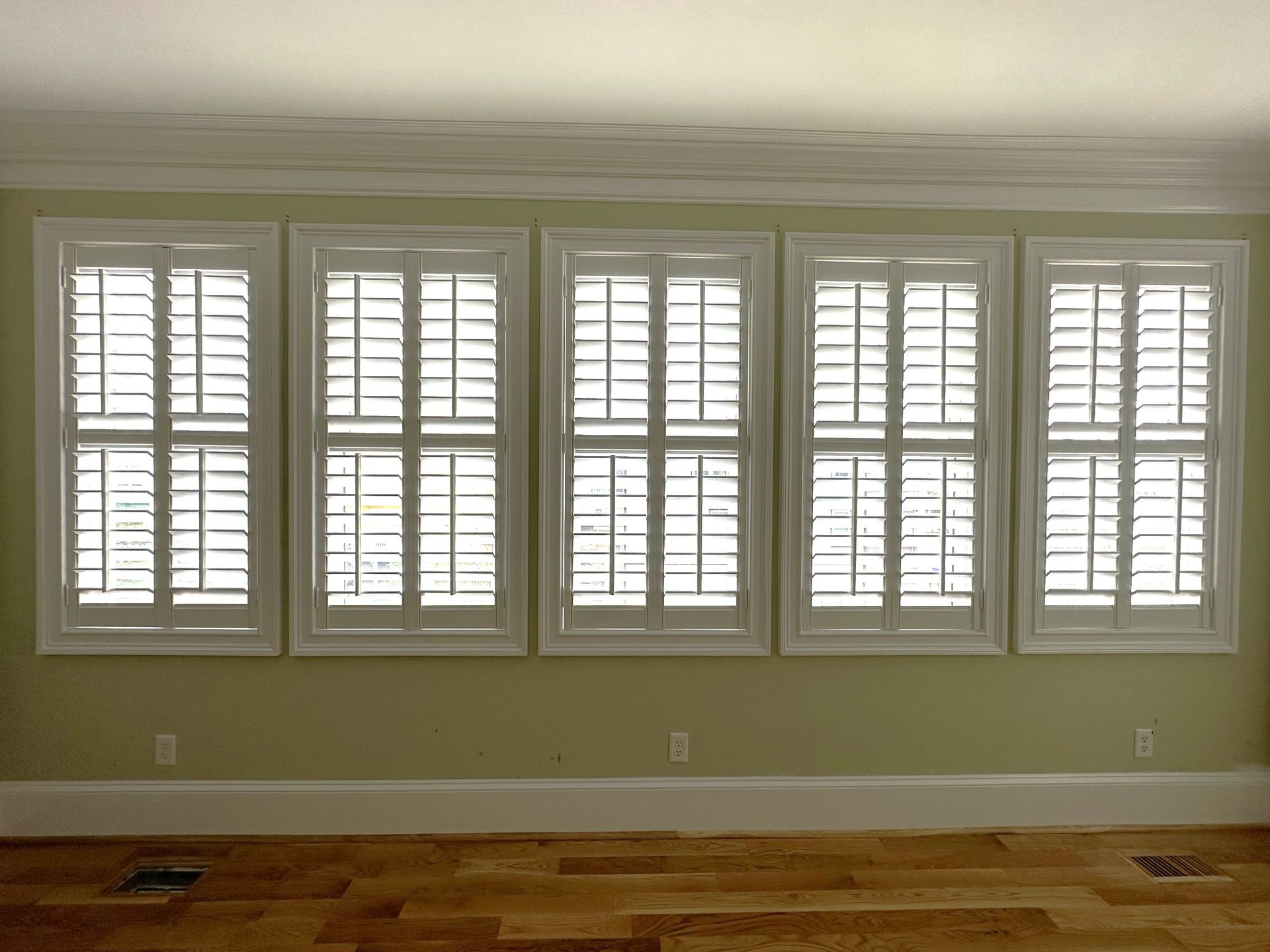 Five white window shutters in a row on a green wall with hardwood floors.