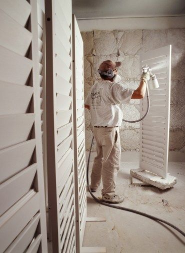 Man sprays white paint on shutters, wearing a mask, inside a room with stone walls.