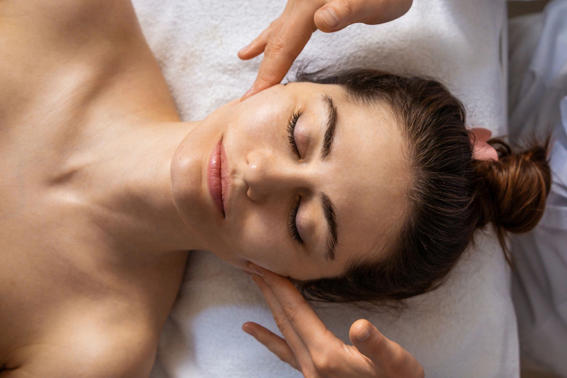 Donna che riceve un massaggio al viso con le mani sulle guance, sdraiata su un asciugamano bianco.