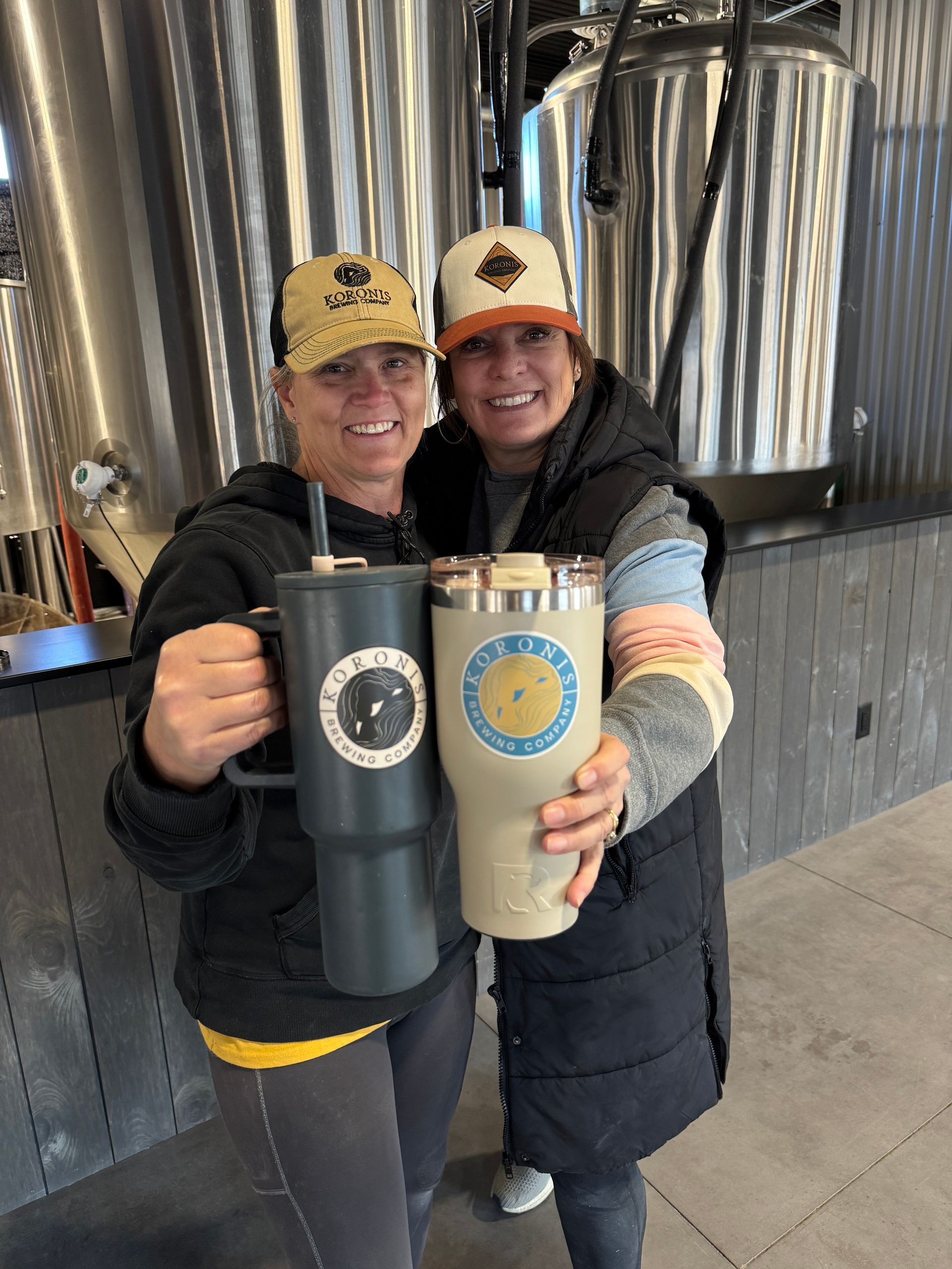 A close up of two women wearing KBC hats and holding KBC mugs merch