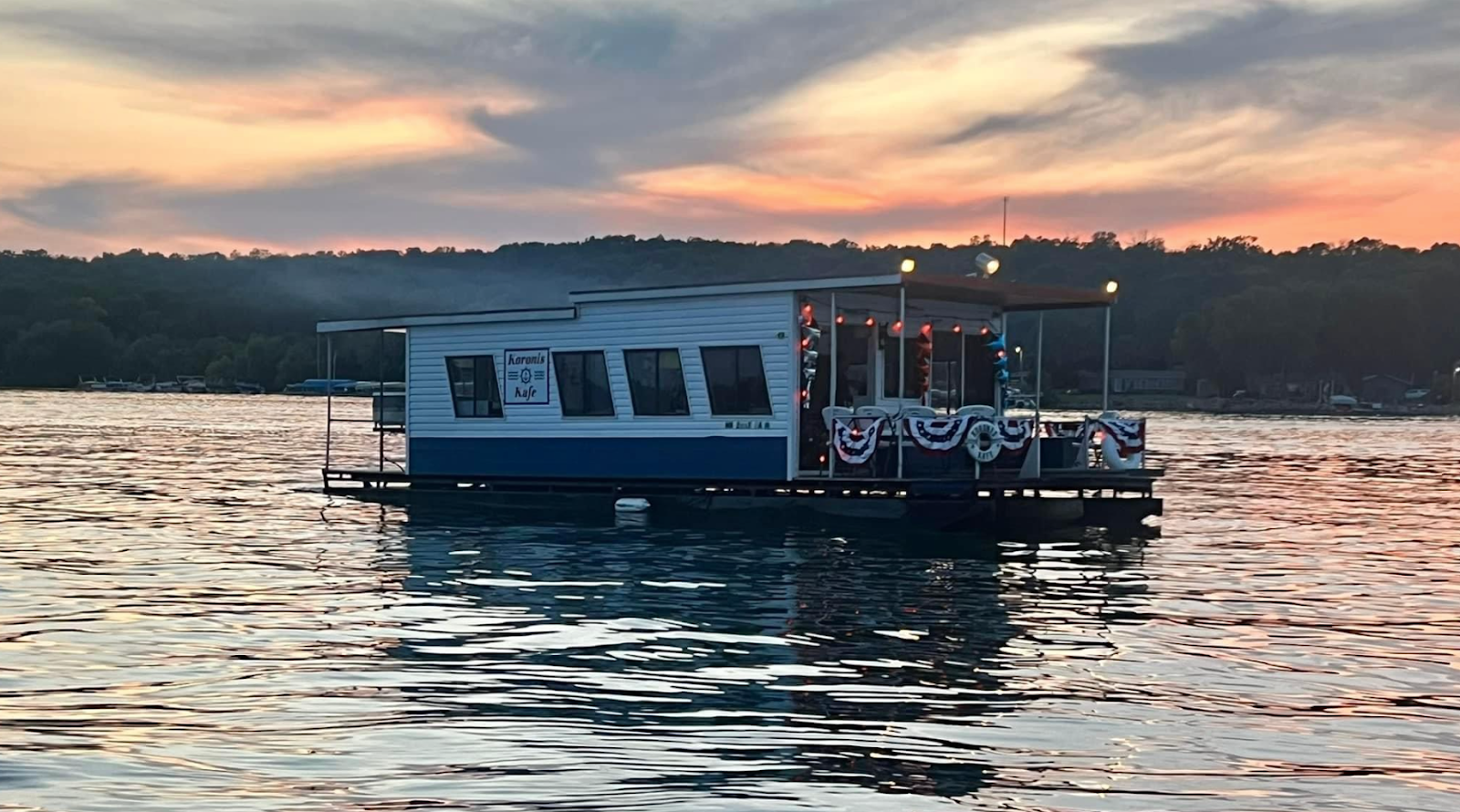 Houseboat with lit deck floating on calm water at sunset, silhouetted shoreline in the background