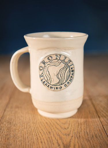 White ceramic mug with a handle and circular logo on a wooden table against a blue background