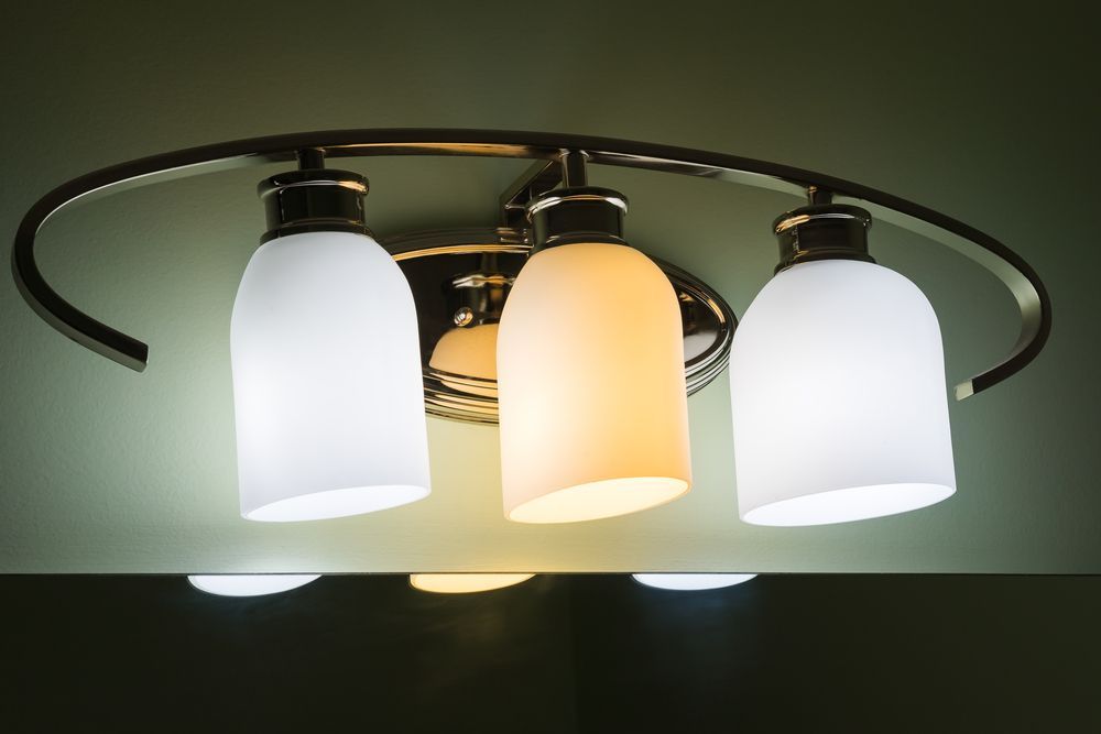 Three-bulb bathroom light fixture with two cool and one warm-toned glowing bulbs on a green wall.