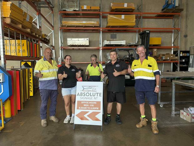Five people giving thumbs up in a warehouse with road signs. They stand behind a sign for