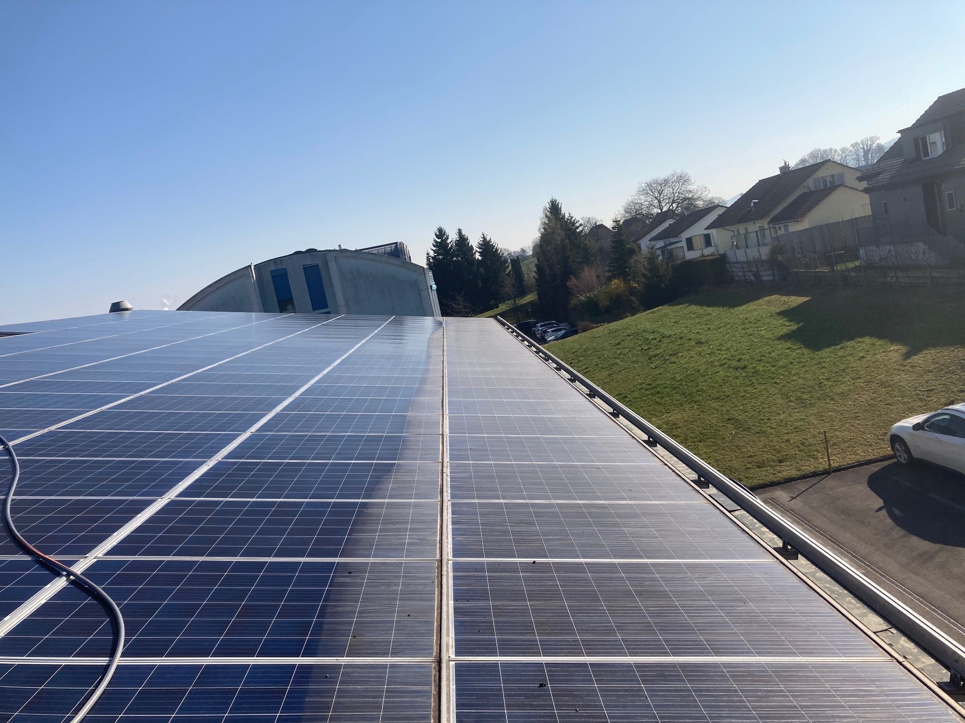 Solar panels on a rooftop beside a grassy hillside under a clear blue sky.
