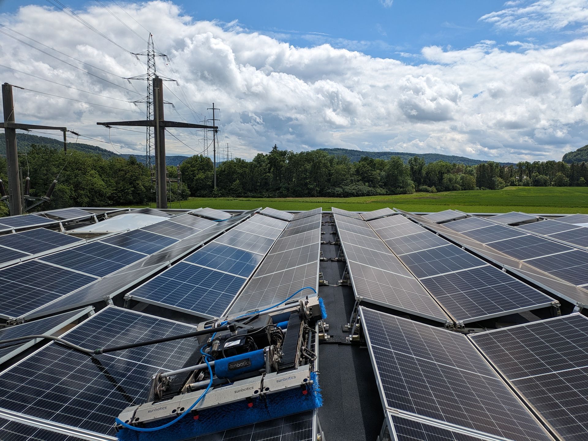 A cleaning robot brushes solar panels on a roof, with power lines and green fields in the background.