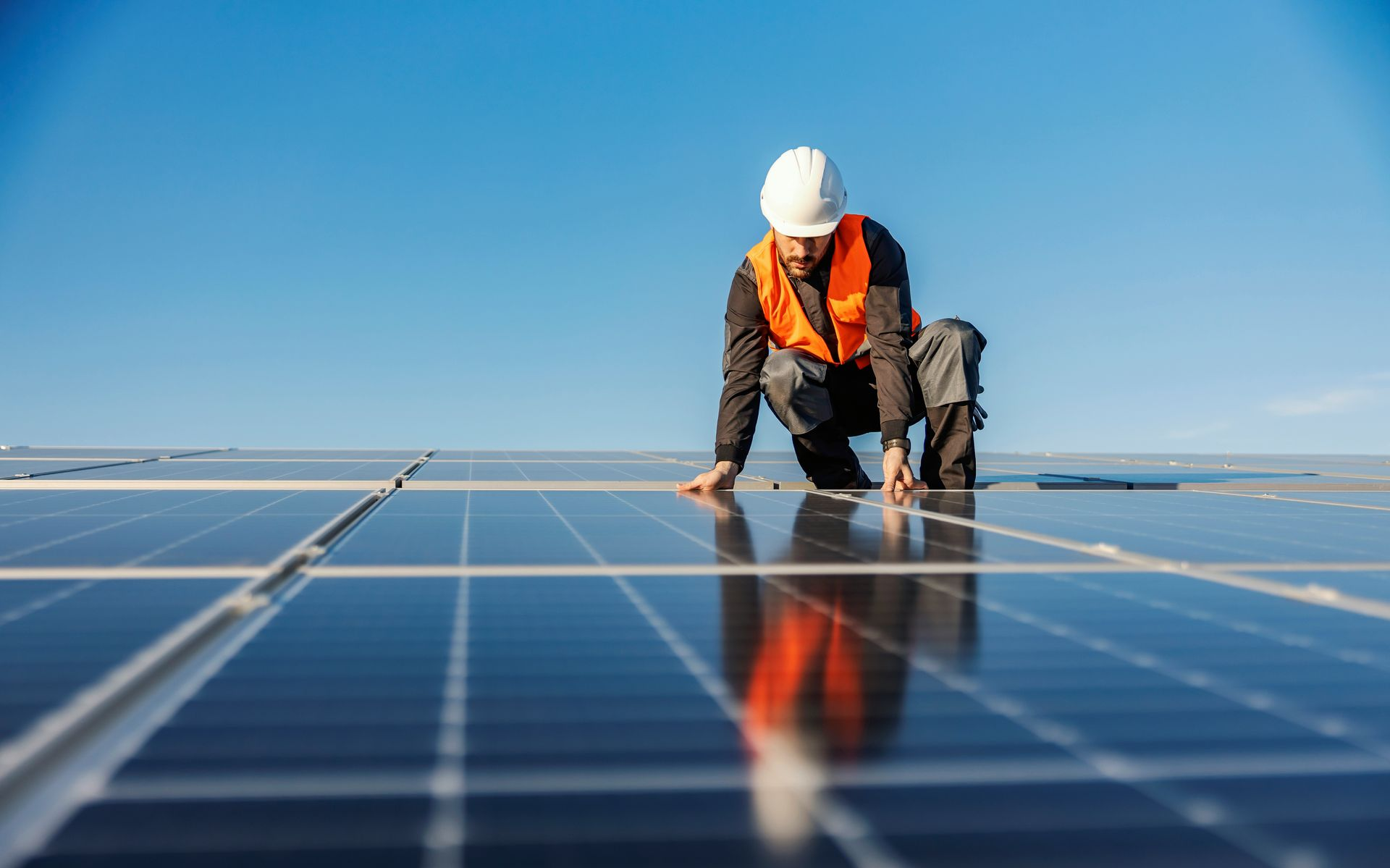 Ein Arbeiter mit Schutzhelm kniet unter einem klaren blauen Himmel auf Solarmodulen und inspiziert sie.
