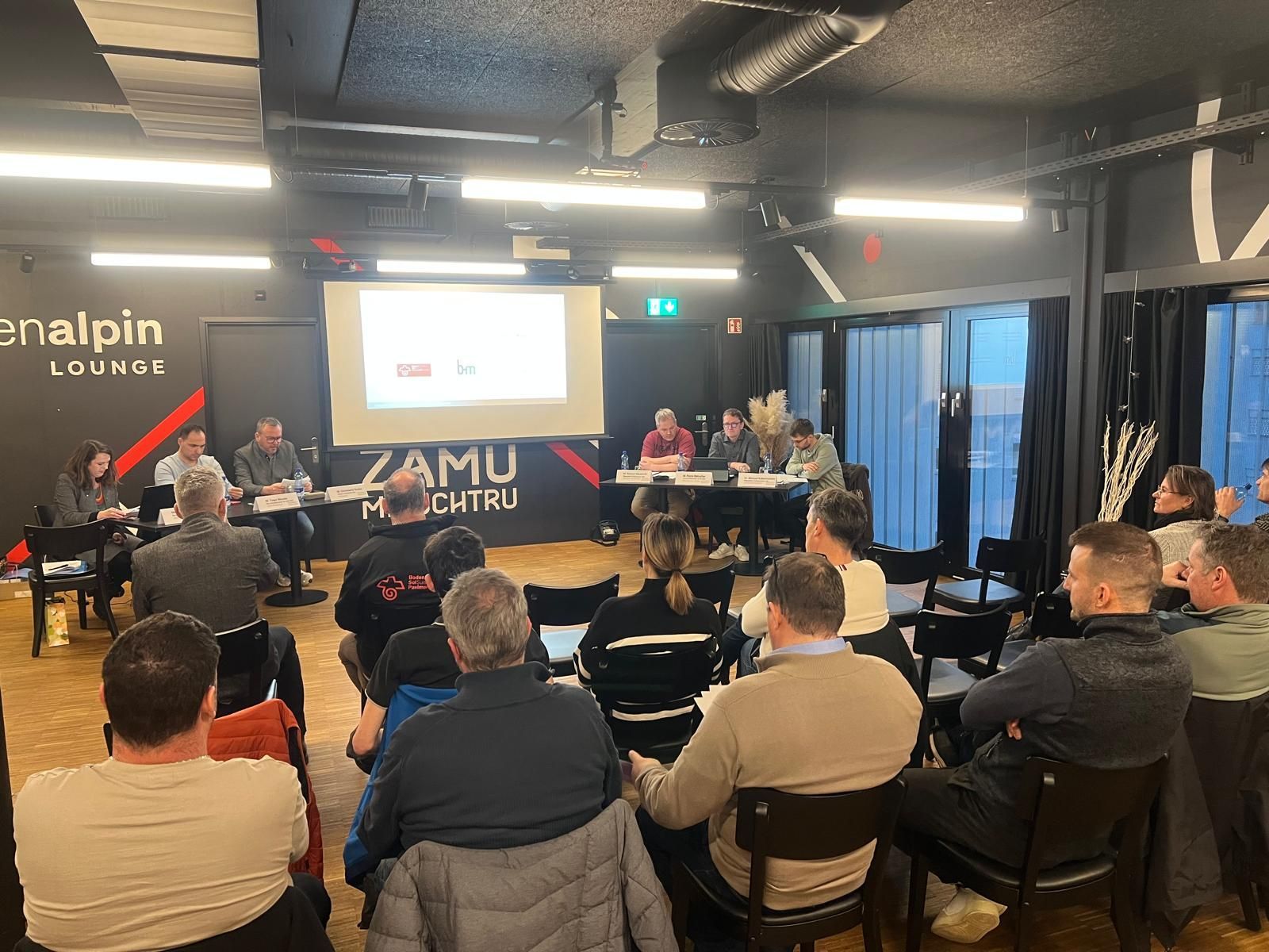 Le public, assis dans un salon lors d'une conférence technique, fait face à une diapositive projetée et aux intervenants, le tout baigné de lumière vive.