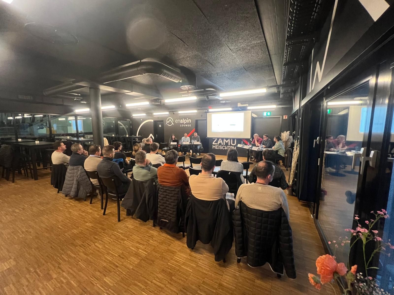 Le public est assis à des tables de banquet face à un écran de présentation dans une salle intérieure moderne.