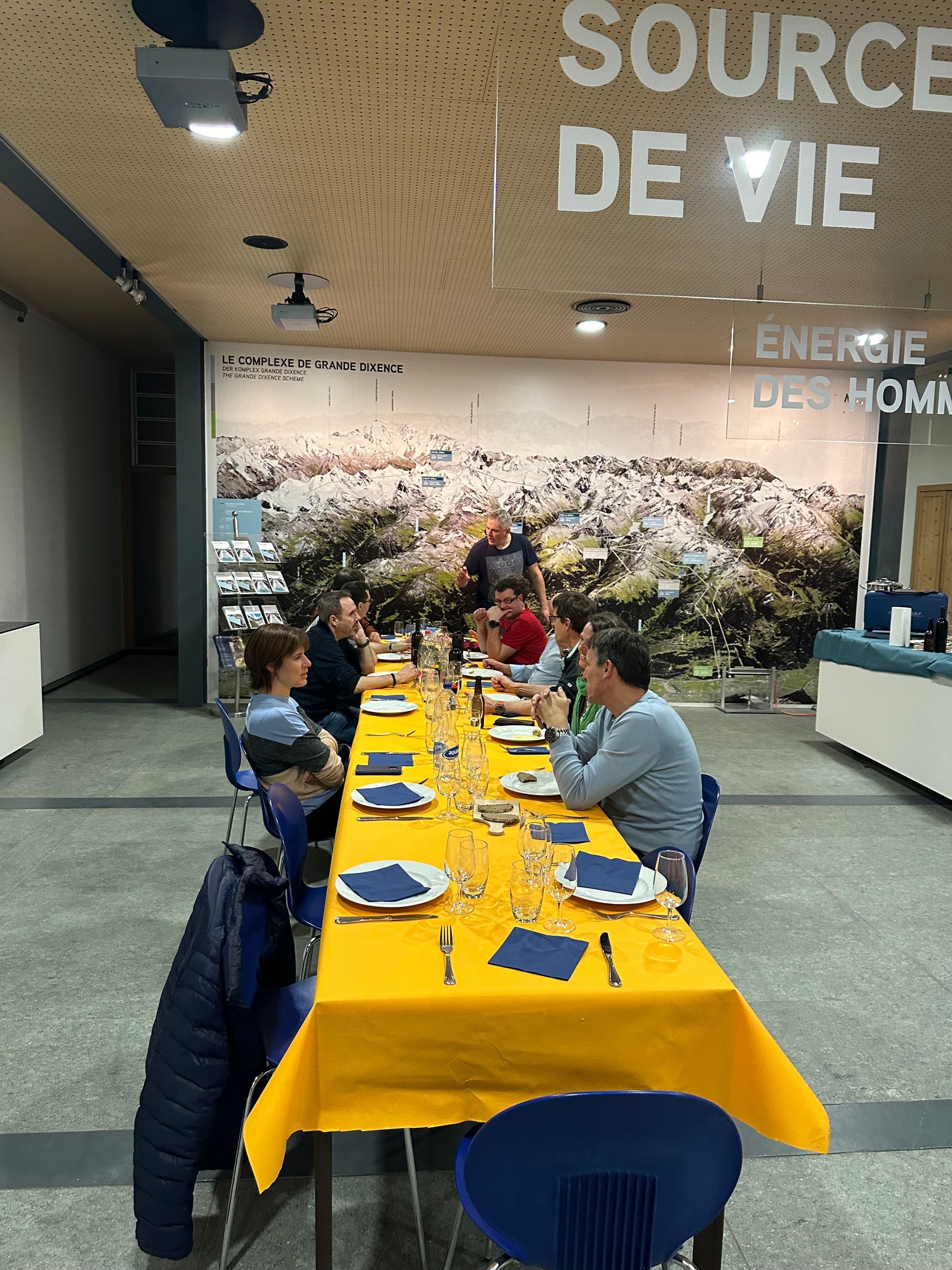 Eine Gruppe von Menschen sitzt an einem langen Tisch vor einer Wand mit der Aufschrift „Source de vie“