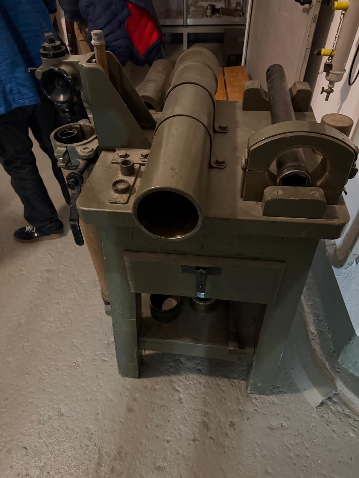 Vieille machine en métal vert dans un atelier, avec un gros tuyau cylindrique et une manivelle sur le dessus.
