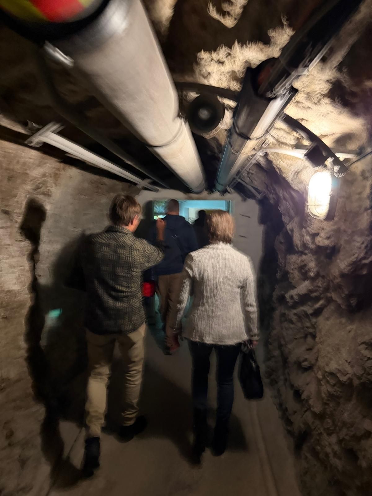 Des personnes traversent un étroit tunnel souterrain avec des tuyaux le long du plafond.