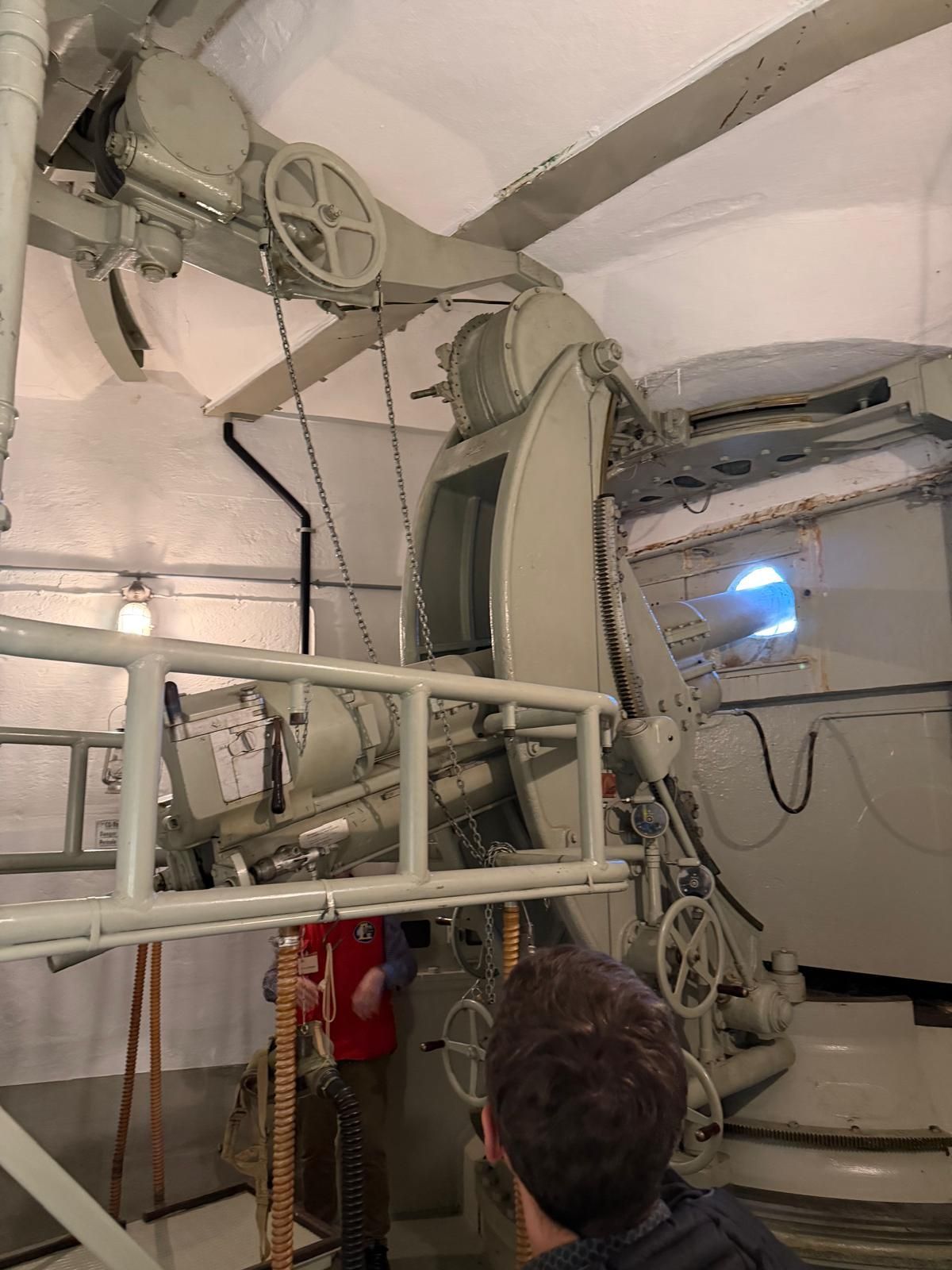 Intérieur d'une grande machine industrielle avec une personne debout en dessous, regardant les câbles et la structure métallique.