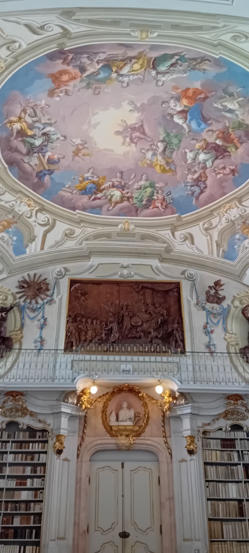 Intérieur de bibliothèque orné, avec un plafond à fresque aux tons pastel, des étagères et une horloge dorée au-dessus de hautes portes blanches.
