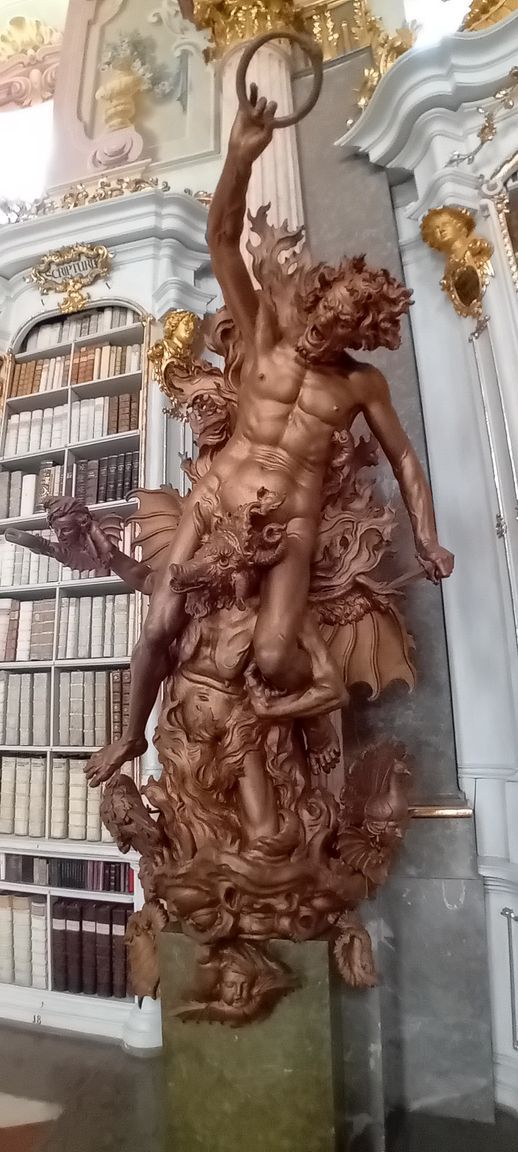Statue en bois sculpté ornée, dans un intérieur décoré avec des étagères et des détails dorés.