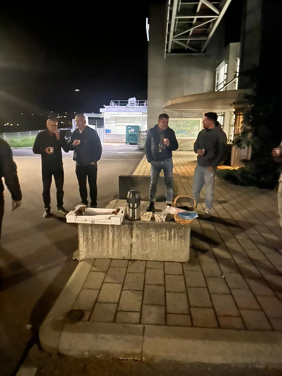 Quatre personnes se tiennent debout sur un trottoir, de nuit, à côté d'un banc bas en béton, avec du matériel et des sacs.