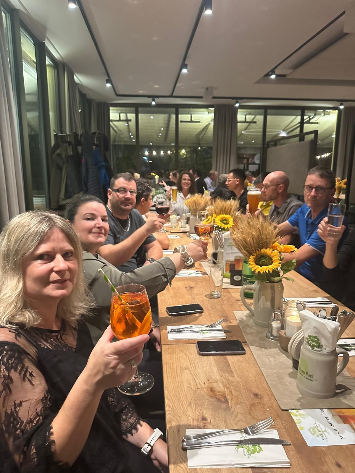 Repas de groupe à une longue table avec boissons et tournesols dans un restaurant lumineux aux parois de verre