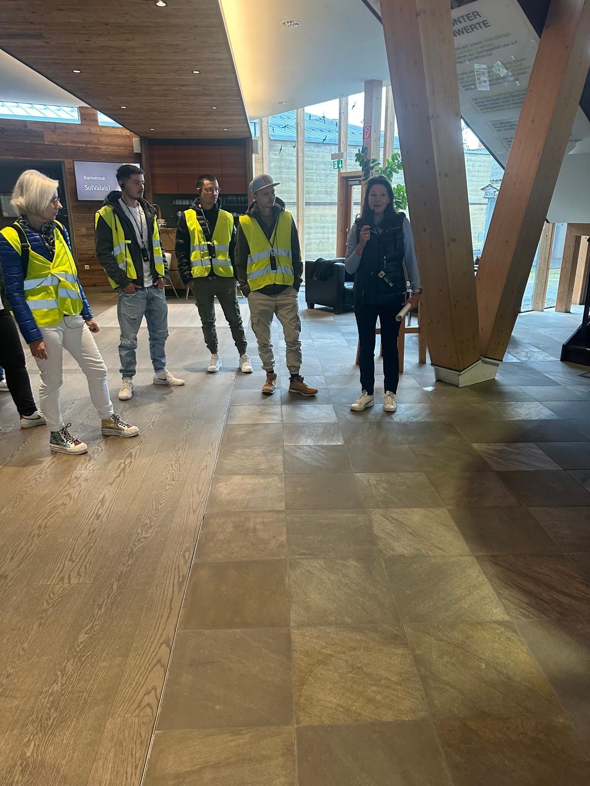 Cinq personnes portant des gilets de sécurité jaunes traversent un hall d'entrée intérieur moderne.