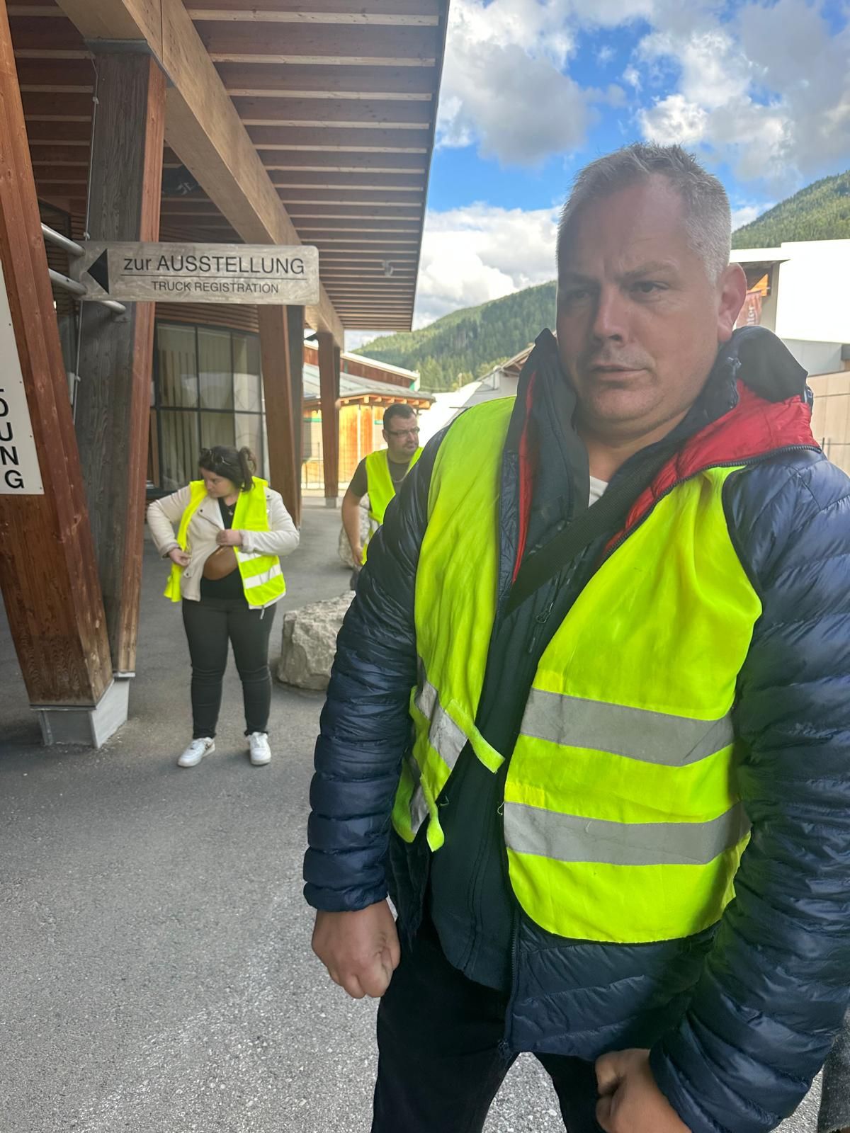 Deux personnes portant des gilets de sécurité fluo se tiennent devant un bâtiment en bois, sur un sentier de montagne ensoleillé.