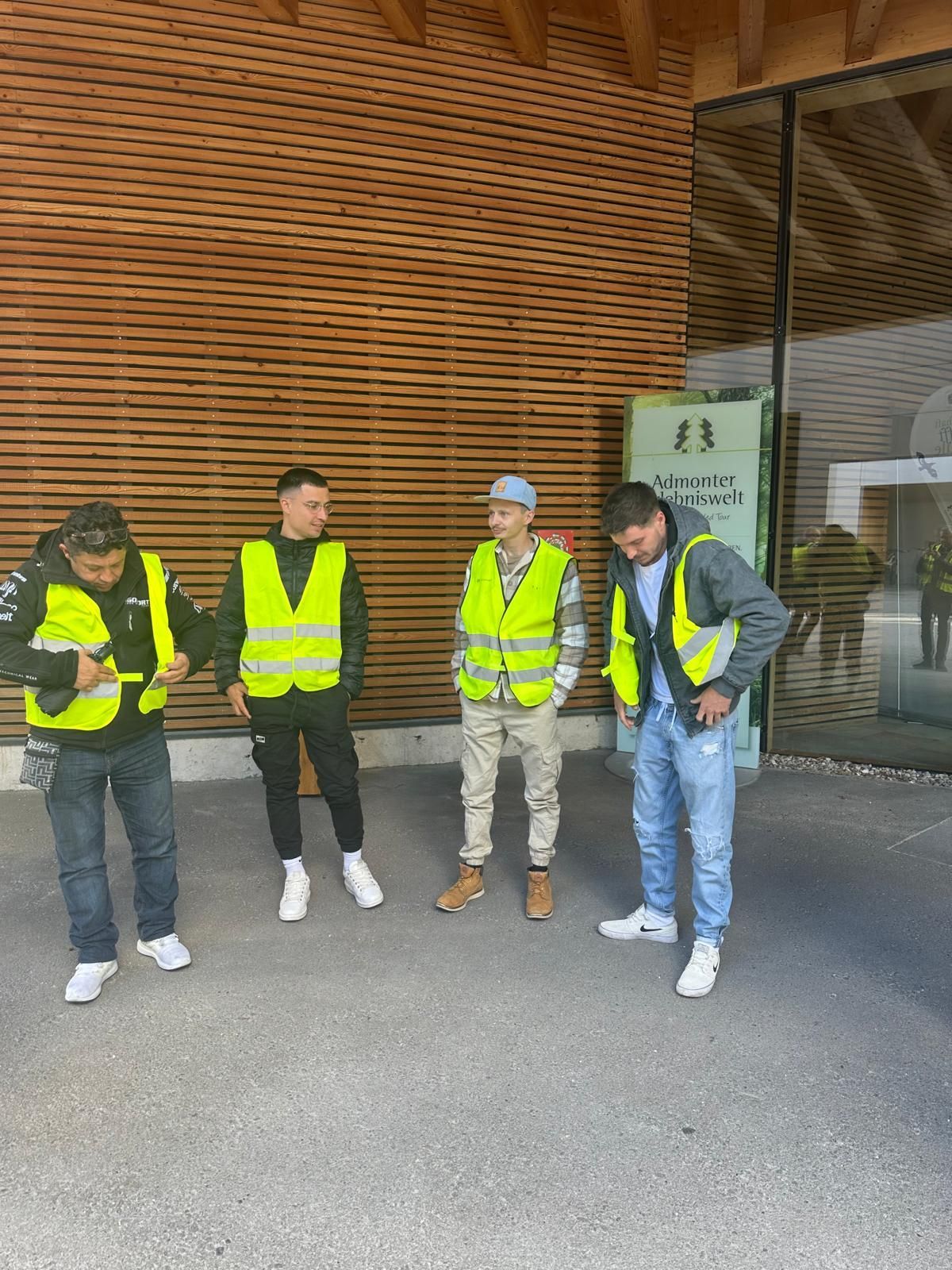 Quatre ouvriers en gilets de sécurité jaunes se tiennent devant un bâtiment en bois, l'un d'eux porte un casque de chantier.
