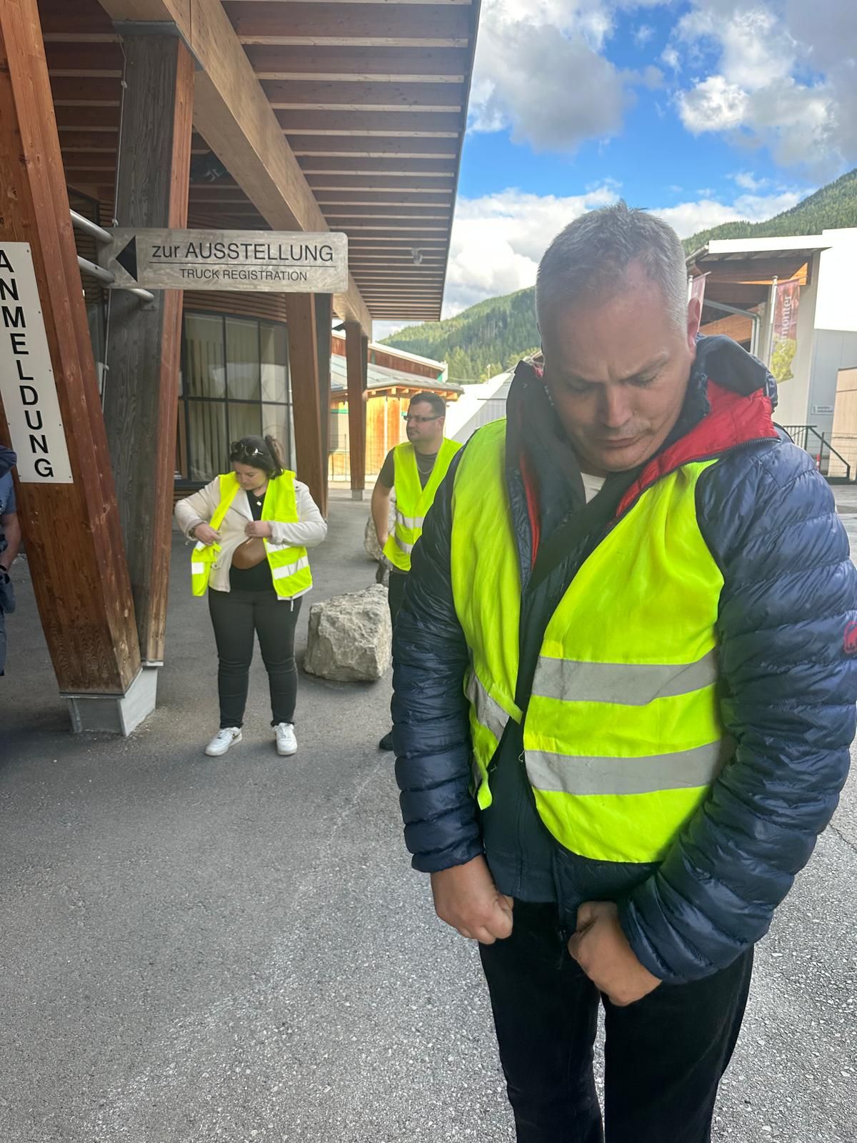 Des personnes portant des gilets de sécurité fluo se tiennent devant un bâtiment en bois ; l'une d'elles ajuste sa veste au premier plan.