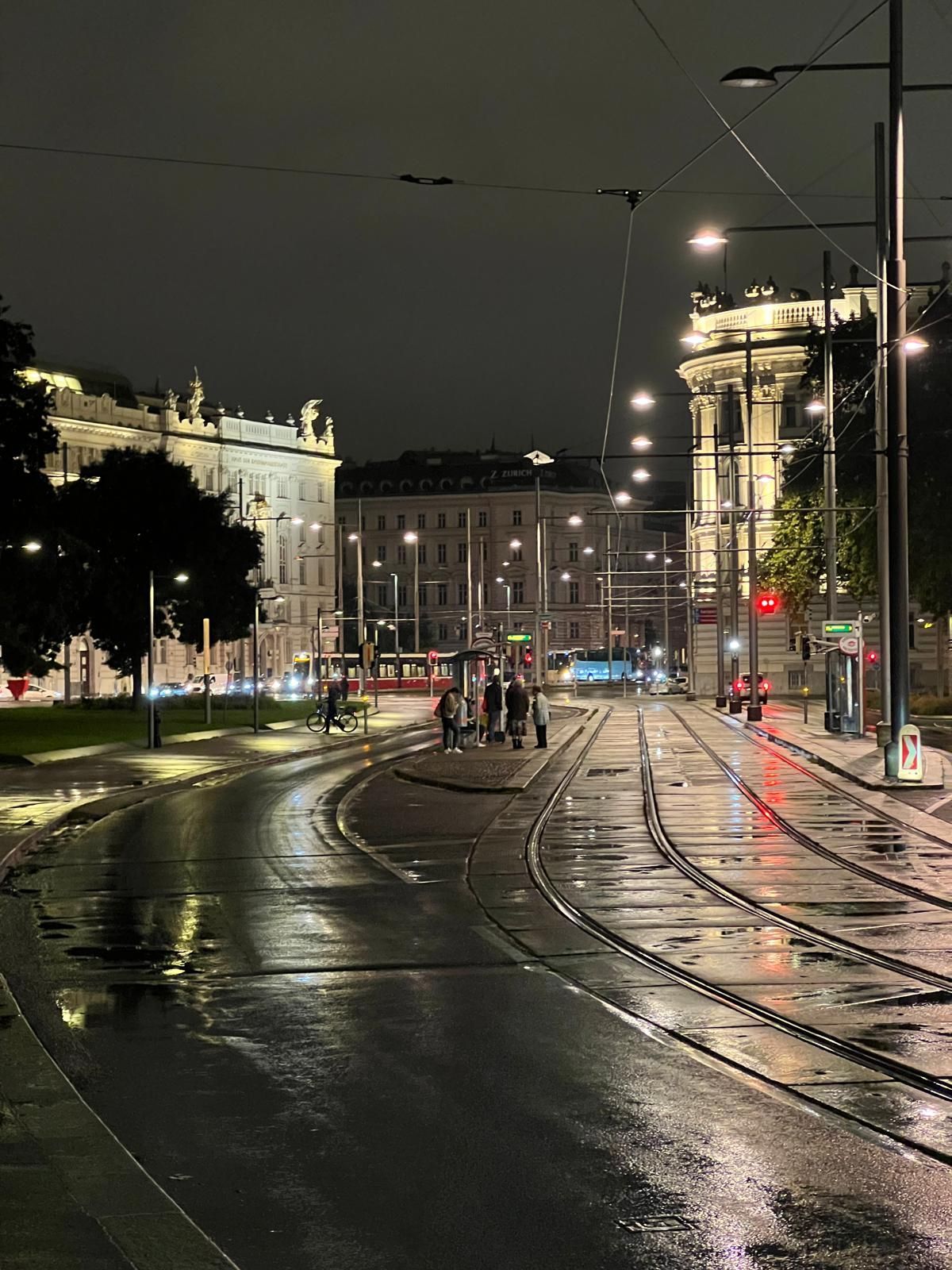 Rue de ville sous la pluie, les rails de tramway et les bâtiments illuminés se reflétant sur le trottoir mouillé.