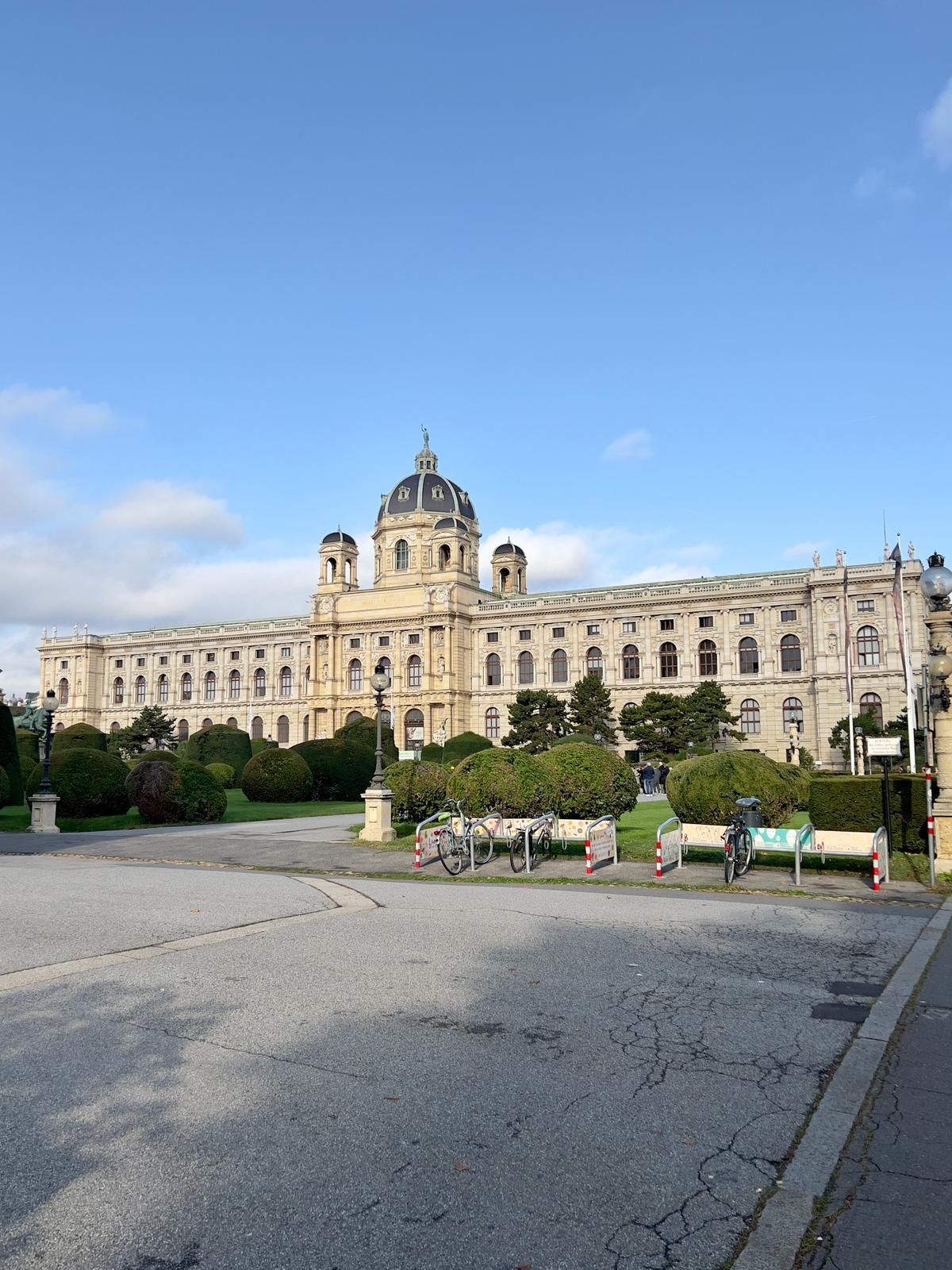 Grand bâtiment historique richement orné, surmonté d'un dôme central, vu depuis une place pavée sous un ciel bleu.