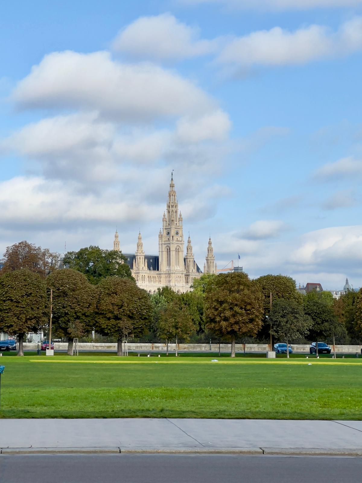 Une vaste pelouse verdoyante bordée d'arbres s'étend devant une haute cathédrale gothique sous un ciel partiellement nuageux.