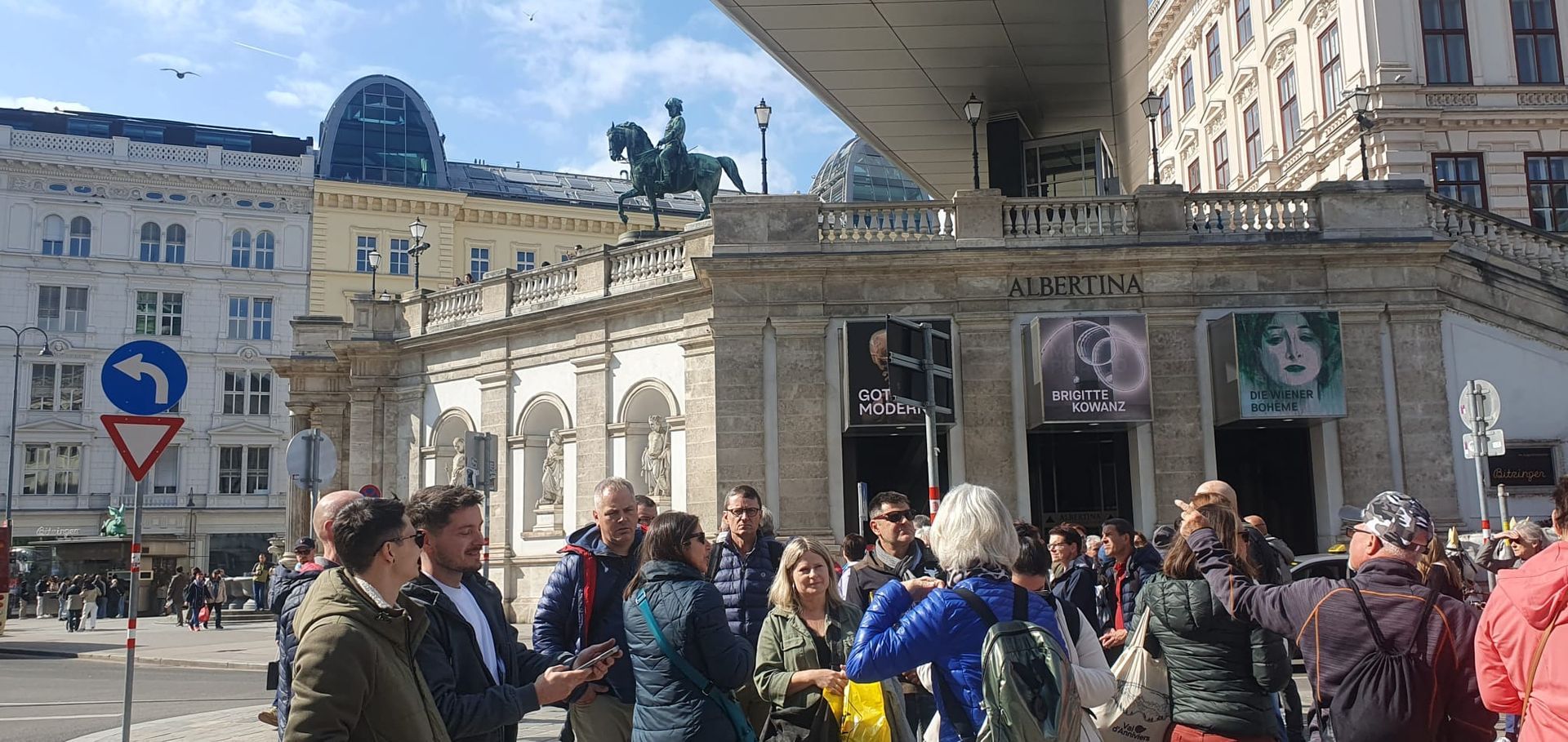 Foule de gens sur une place de la ville, près d'un bâtiment en pierre orné de statues et de drapeaux, sous un soleil éclatant.