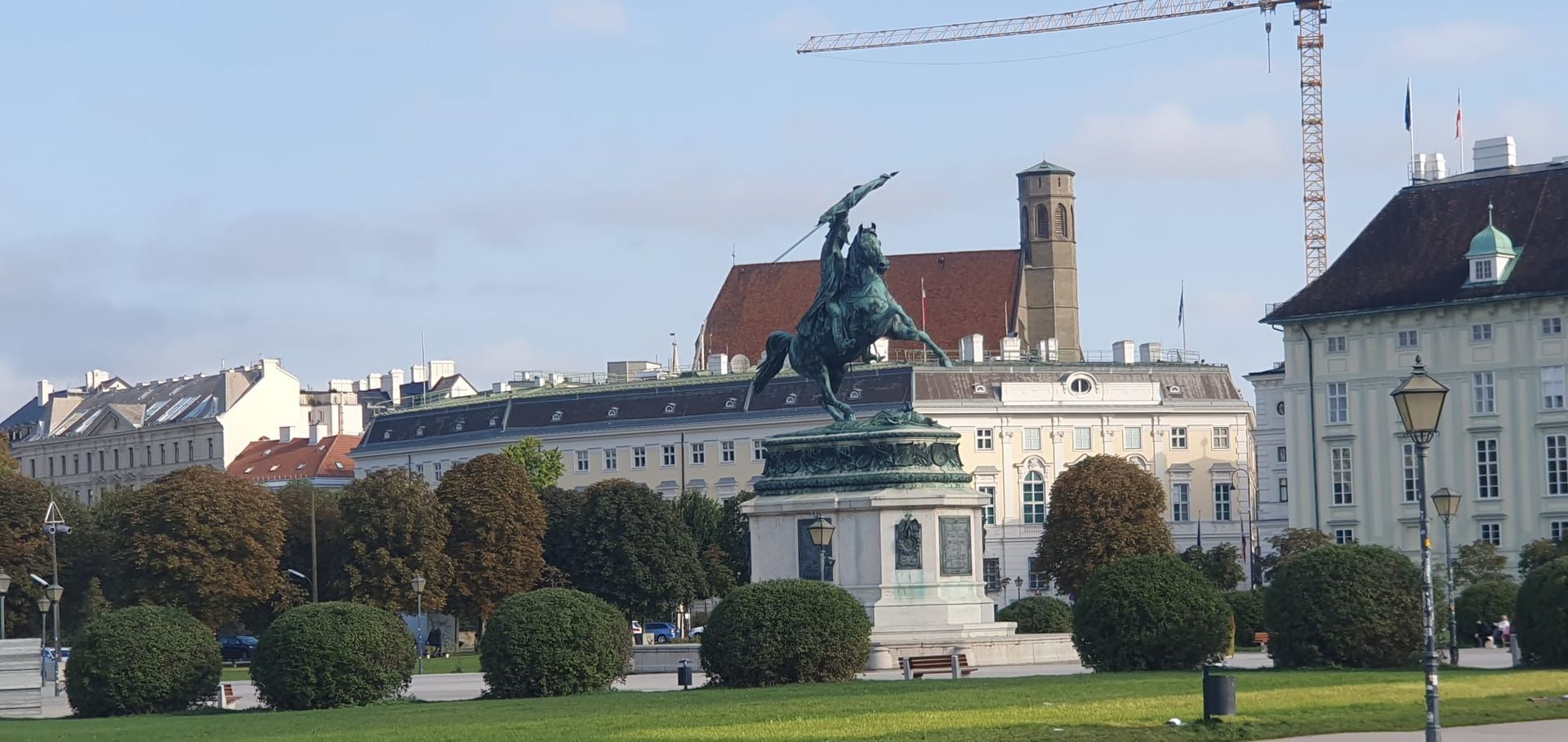 Statue sur une place européenne avec pelouses impeccables, bâtiments historiques et une grue en arrière-plan