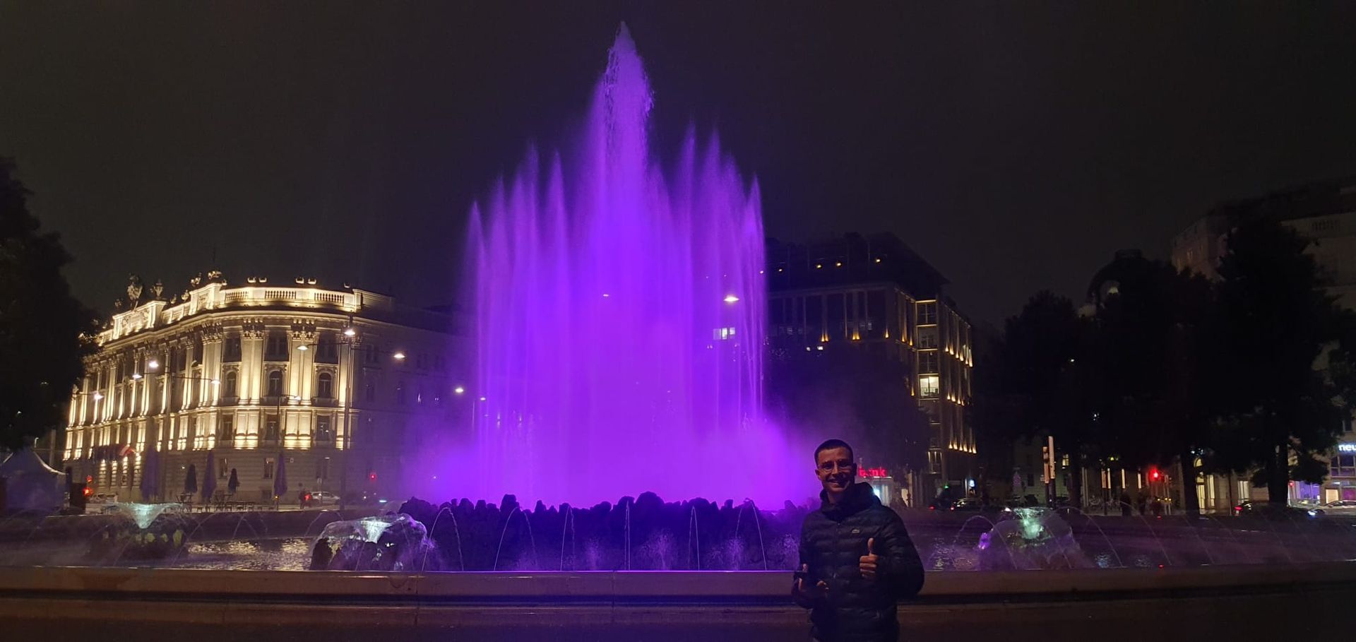 Fontaine nocturne illuminée de violet devant un bâtiment éclairé, une personne prenant une photo au premier plan