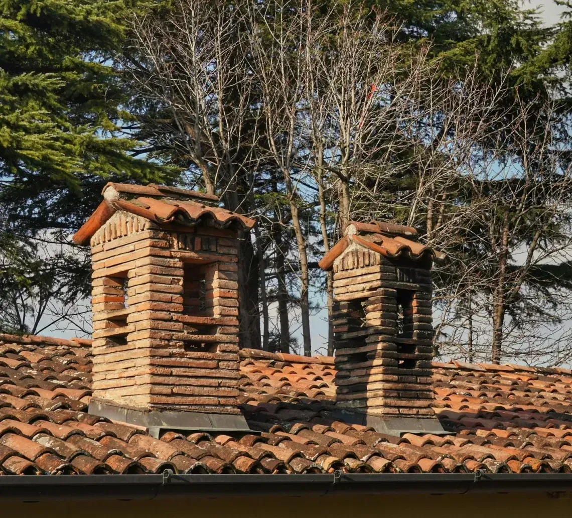 Two brick chimneys on a terracotta tile roof with tall trees in the background