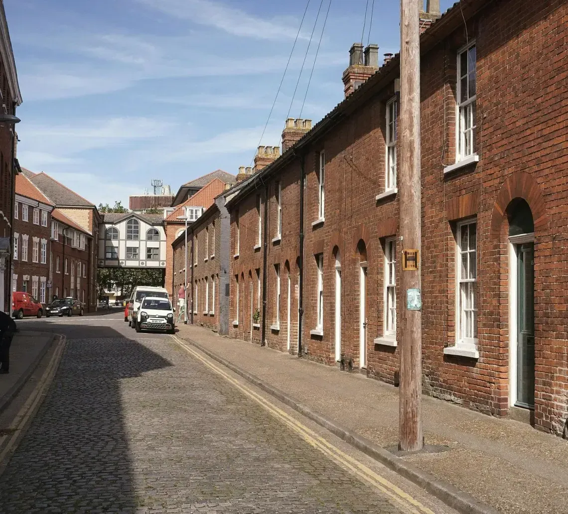 Narrow cobblestone street lined with red brick houses and parked cars under a blue sky
