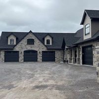 Large stone house with three garage doors and a wide gravel driveway under a cloudy sky