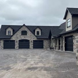 Large gray stone house with multiple garage doors and a wide driveway under cloudy skies