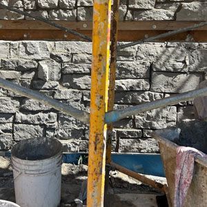 Yellow rusty metal post with crossed supports in front of a stone wall and bucket