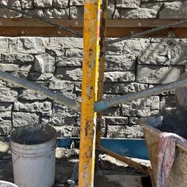 Rusty yellow metal scaffold against a stone wall, with a white bucket and cloth nearby.