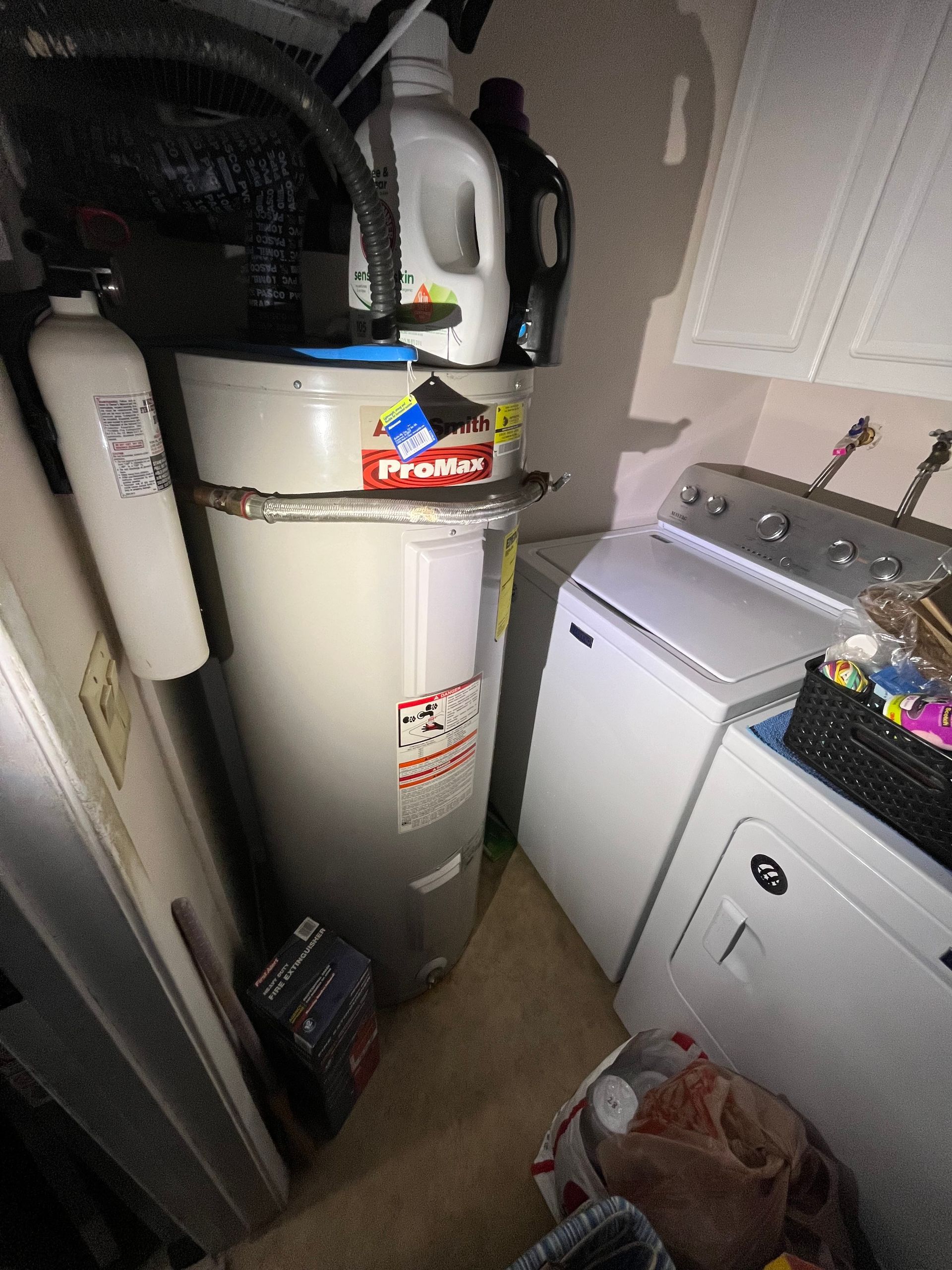 Water heater beside a laundry machine in a cramped utility closet, with cleaning supplies on top.