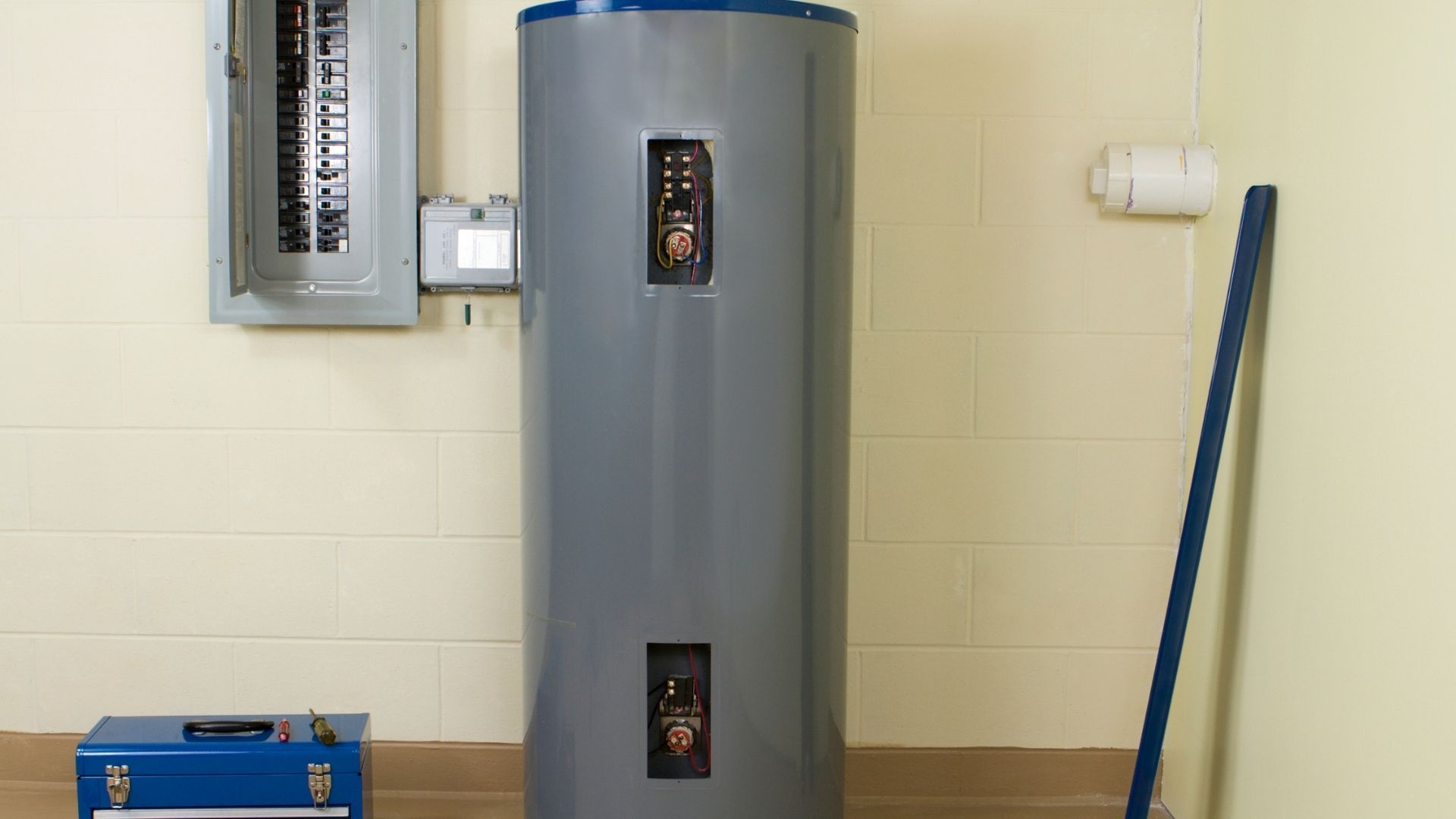Water heater in a utility room with an electrical panel, blue toolbox, and broom along the wall.