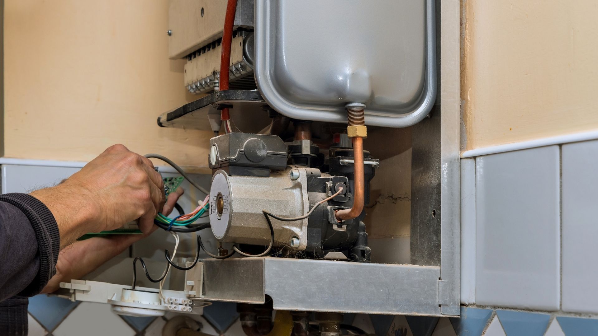 Hand adjusting wiring on a boiler unit with copper pipes and a silver expansion tank.