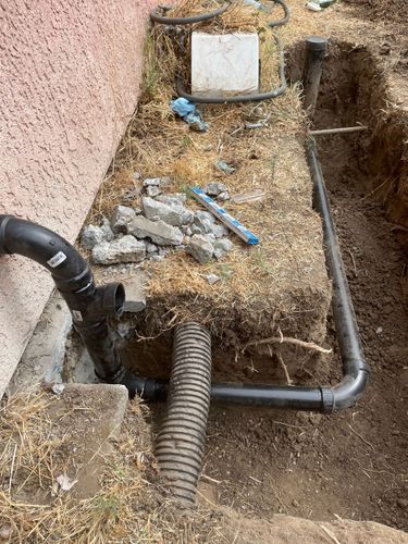 Damaged outdoor utility area with exposed pipes, dirt, gravel, and a broken hose near a wall.