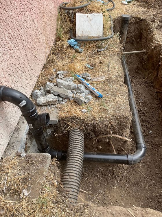 Damaged outdoor utility area with exposed pipes, dirt, gravel, and a broken hose near a wall.