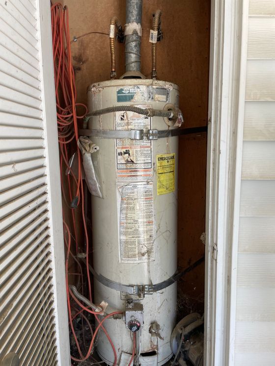 Water heater in a narrow utility closet with pipes, labels, and red cords along the left wall.
