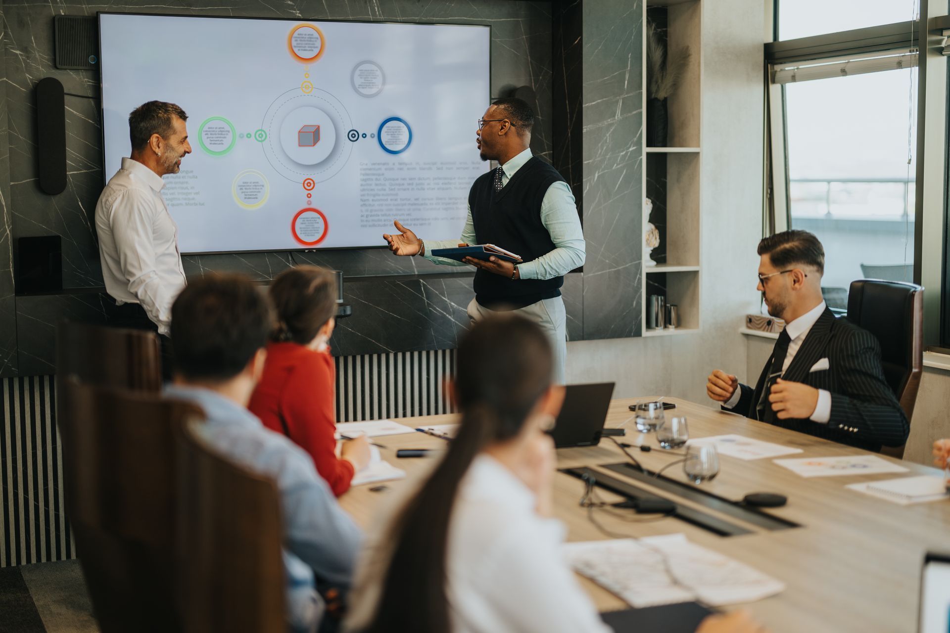 Business meeting with a presenter pointing at a wall screen while colleagues listen around a conference table Business meeting with a presenter pointing at a wall screen while colleagues listen around a conference table