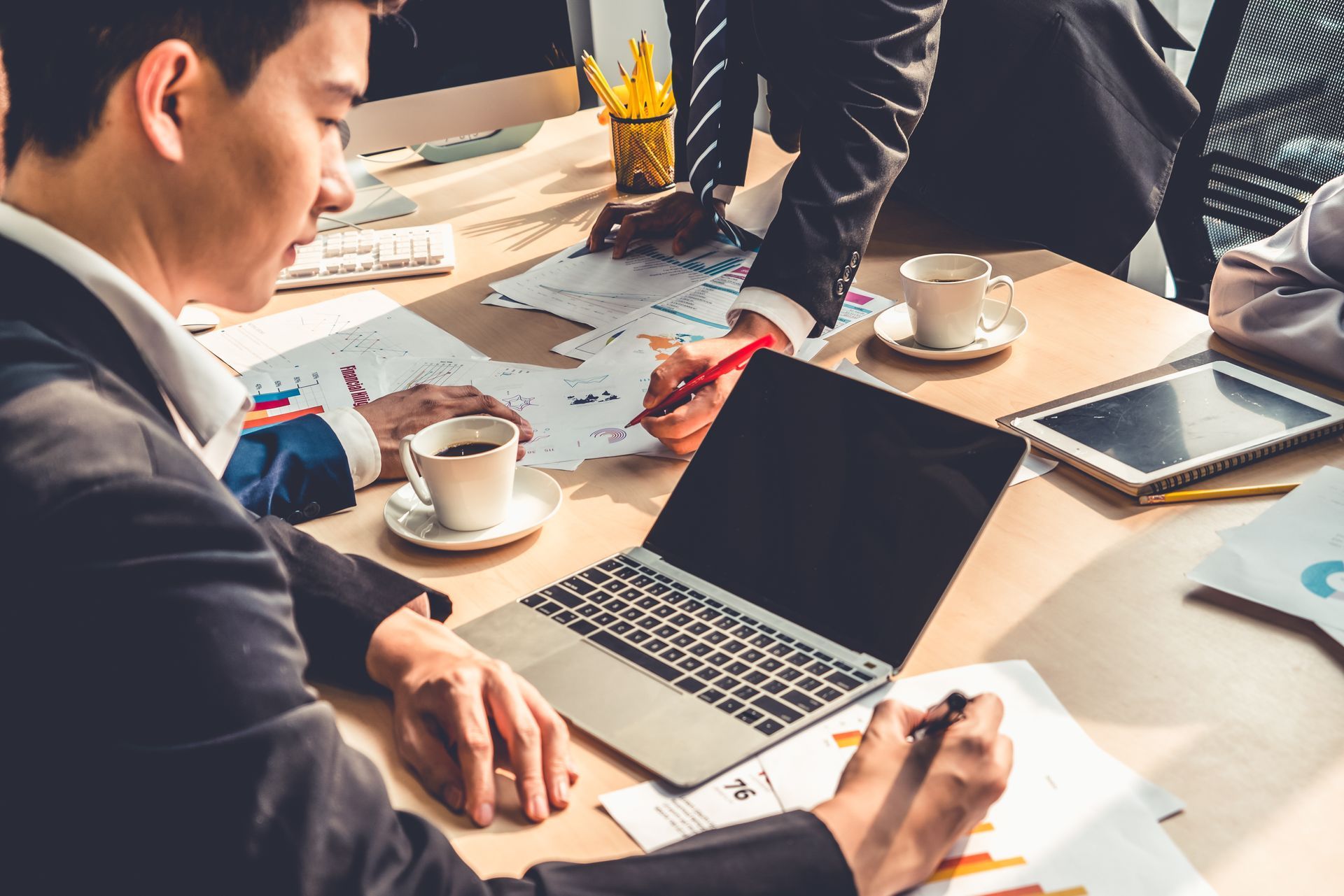 People in a meeting around a laptop, reviewing documents and coffee on a bright conference table. People in a meeting around a laptop, reviewing documents and coffee on a bright conference table.