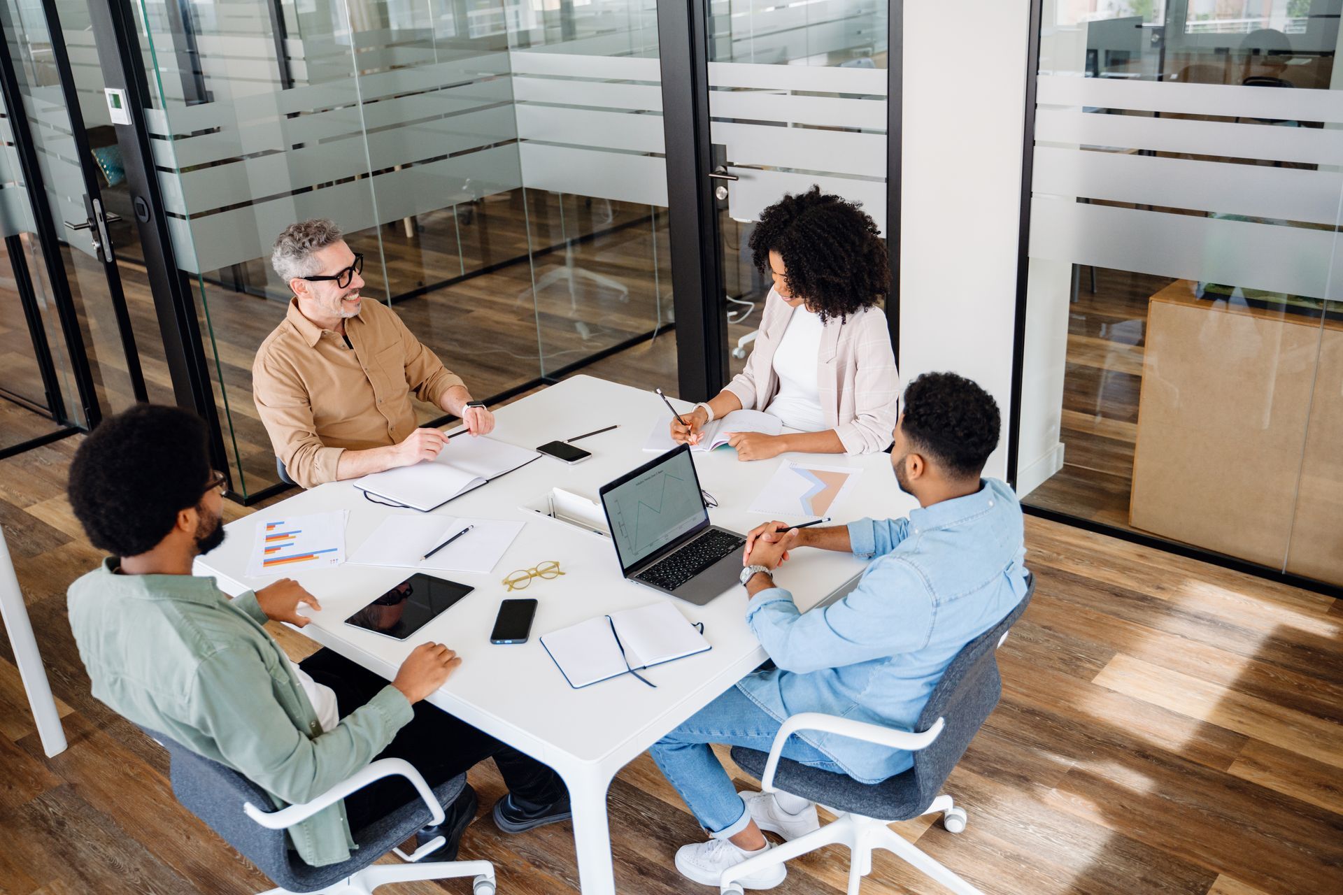 Four coworkers meeting around a table in a glass-walled office, discussing papers and a laptop. Four coworkers meeting around a table in a glass-walled office, discussing papers and a laptop.