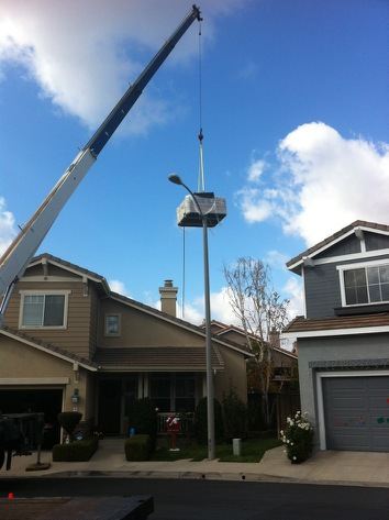 Crane lifting a small house-shaped structure above suburban homes on a sunny day.