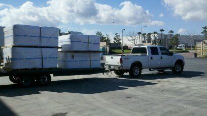 White pickup truck towing a flatbed trailer loaded with stacked white bundles in a parking lot