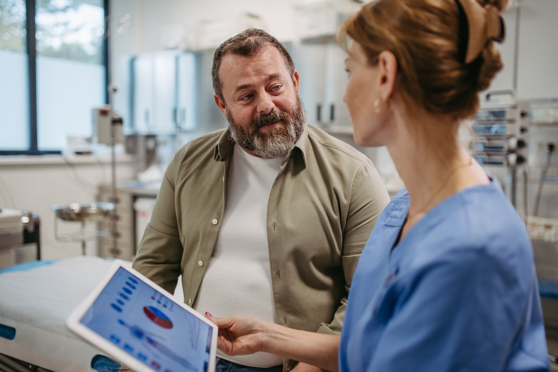 Clinician in scrubs reviews a tablet health chart with a male patient in a medical office Clinician in scrubs reviews a tablet health chart with a male patient in a medical office